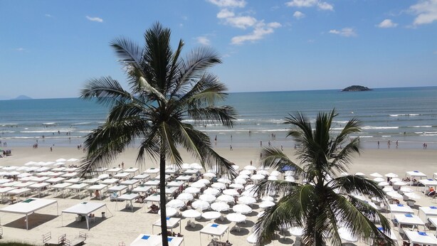A beach with rows of white umbrellas and tables, palm trees, and the ocean. - Excellent 135 m2 apartment 70 m from the sea, with electric barbecue and Wi-Fi  (BERTIOGA)