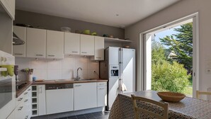Una cocina moderna con armarios blancos, lavavajillas empotrado y refrigerador. Hay una mesa de comedor con sillas y vistas a zonas verdes.