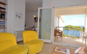 A room with a yellow armchair, a table with a magazine, a bookshelf, and a view of the sea through a balcony door.