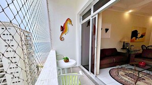 Un balcón con una mesa y sillas blancas, un caballito de mar decorativo, y vistas del interior que incluyen un sofá y un tapete estampado.