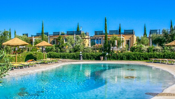 A swimming pool with lounge chairs and umbrellas, surrounded by buildings and greenery.