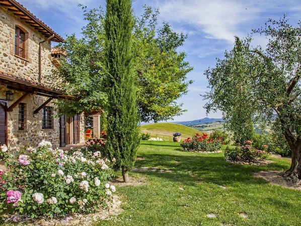 A stone house with a garden, a tall tree, and a view of rolling hills.