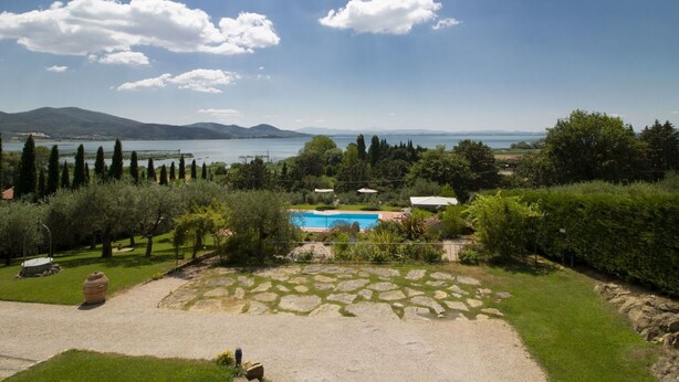 A pool area with stone paving, surrounded by greenery and trees, overlooking a lake and distant mountains.