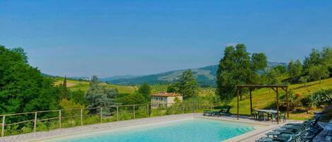 Ein Swimmingpool mit Steinfliesen, umgeben von Liegestühlen und einer Pergola, mit Blick auf eine malerische Landschaft aus Hügeln und Bäumen.