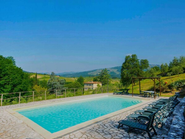 A swimming pool with stone tiles, surrounded by lounge chairs and a pergola, overlooking a scenic landscape with hills and trees.
