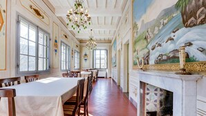 Een lange eetzaal met hoge plafonds, grote ramen en een muurschildering met een landschapsvoorstelling.