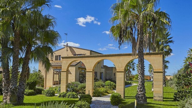 Edifício em estilo mediterrâneo com arcos, cercado por jardins bem cuidados e palmeiras.