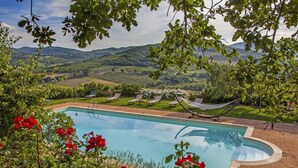 A pool surrounded by lush greenery and red flowers, with a view of distant hills and a clear sky. - Cosy apartment for 6 guests with pool, WIFI, TV, patio, pets allowed and panoramic view (Allerona)