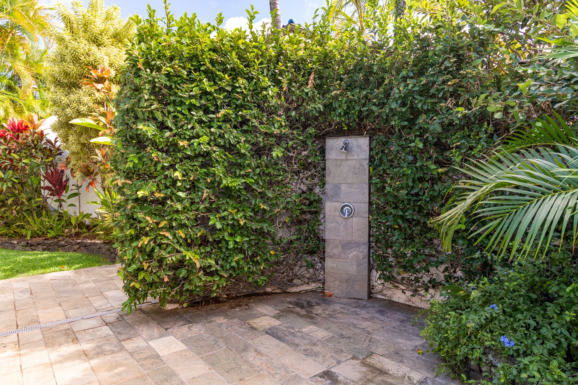 A stone wall with a metal door, surrounded by lush greenery and a paved walkway.