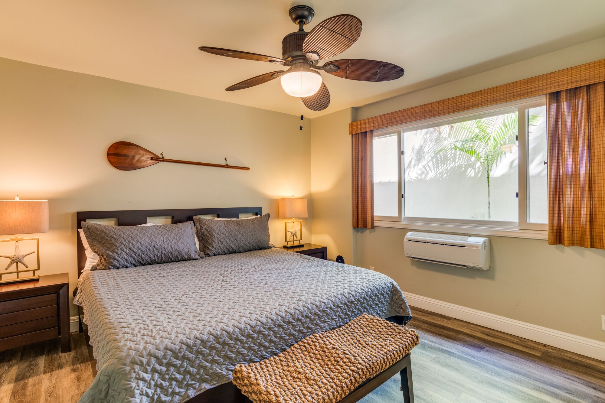 Une chambre à coucher comprenant un lit, des lampes de chevet, un ventilateur de plafond, une fenêtre avec des rideaux et un appareil de climatisation.