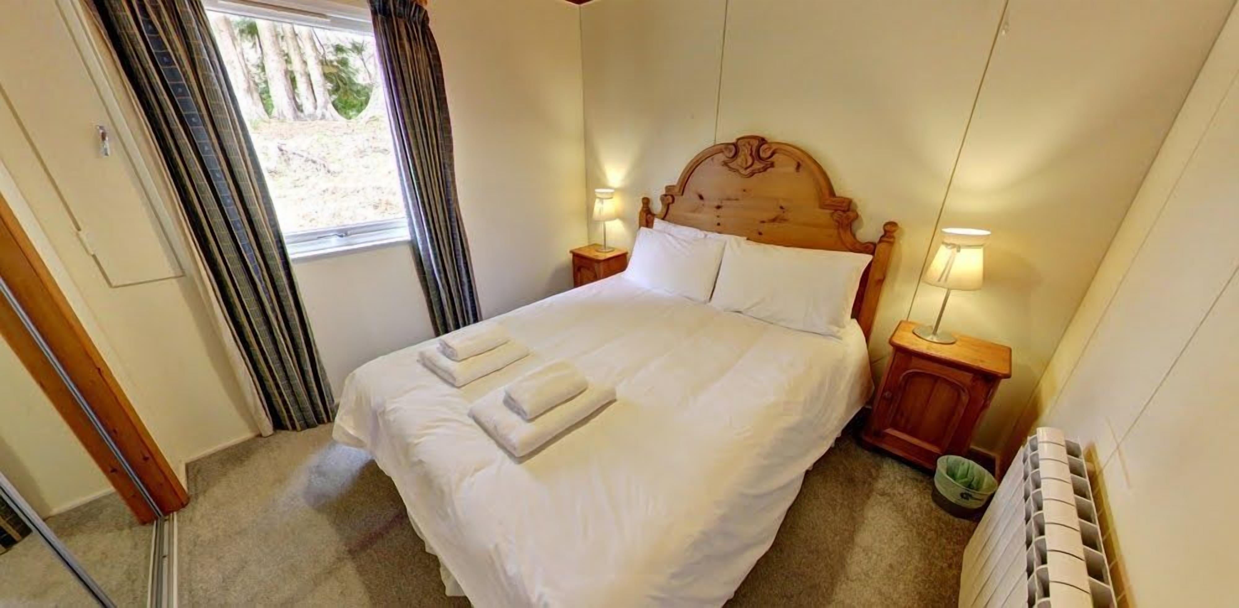 A bedroom with a bed, two bedside lamps, a window with curtains, and a radiator.