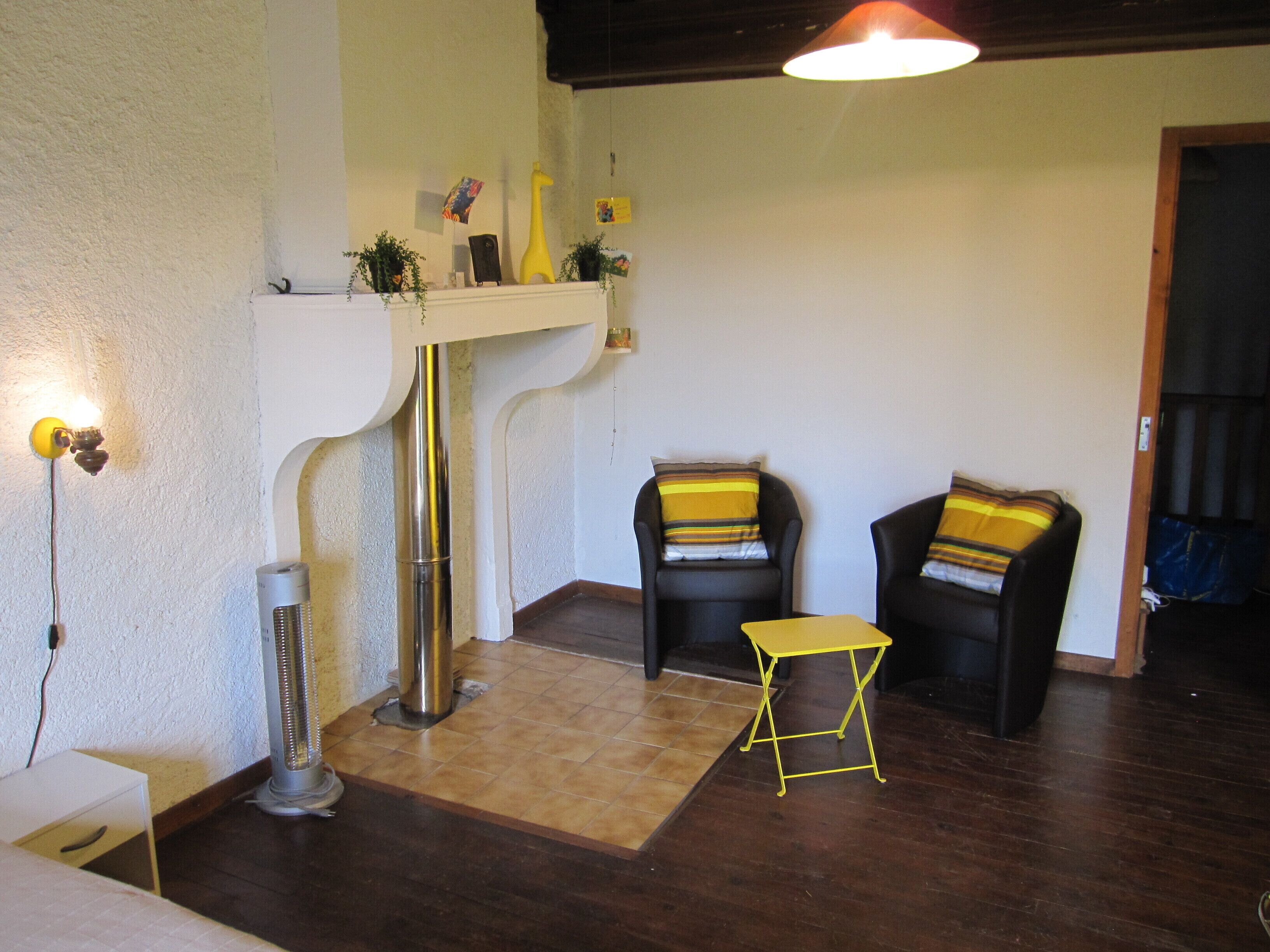 Una habitación con pared blanca, suelo de madera, una mesa amarilla, dos sillas negras con cojines a rayas, un estante blanco con objetos decorativos y un calentador portátil.