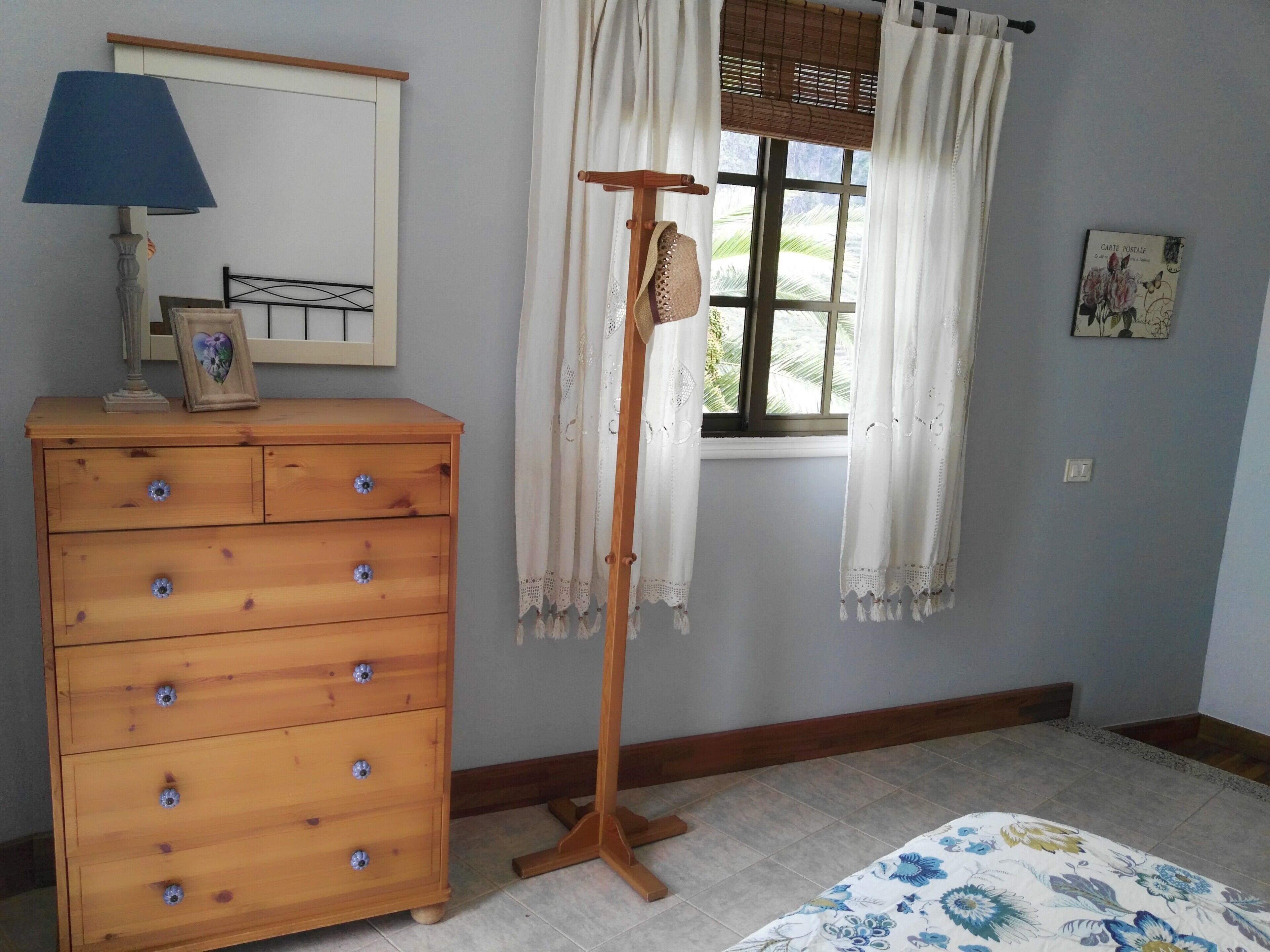 Une chambre à coucher avec une commode en bois, un lampadaire, une fenêtre avec des rideaux et un lit recouvert d’une couvre-lit à motifs floraux.