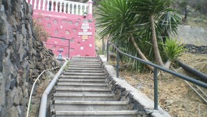 Eine Treppe führt zu einem rosa Gebäude mit Löchern, umgeben von trockenem Gras und Palmen.