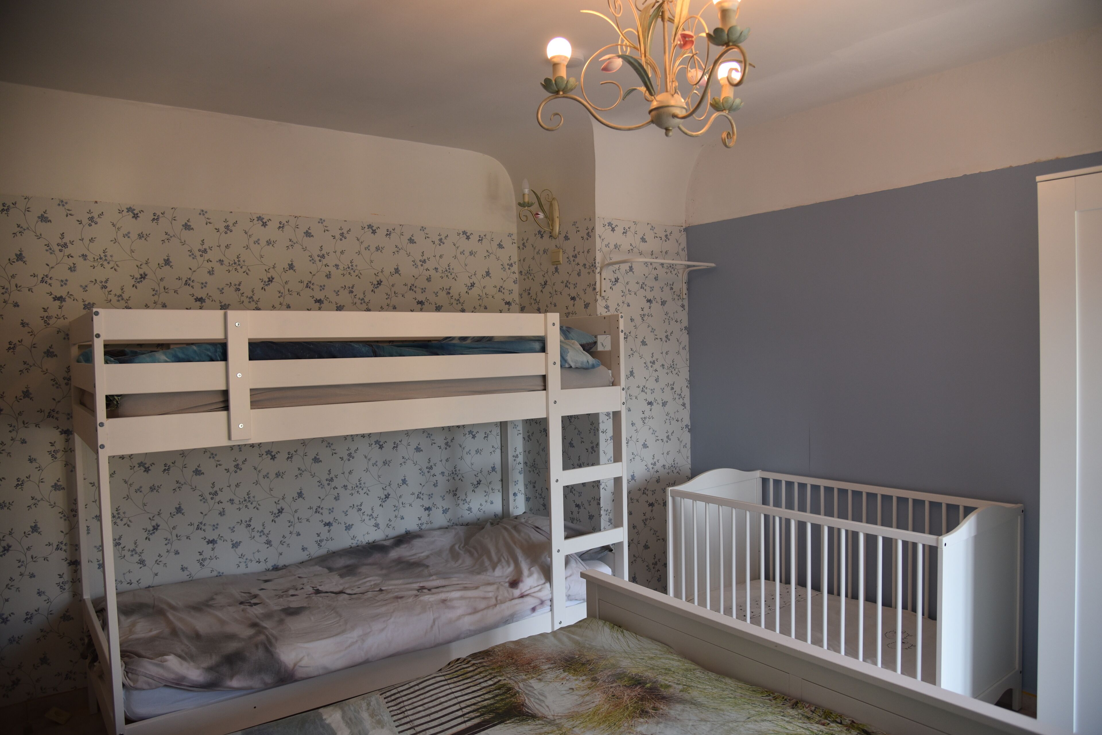 A bunk bed with a crib attached to the right side, a floral wallpaper, and a chandelier.