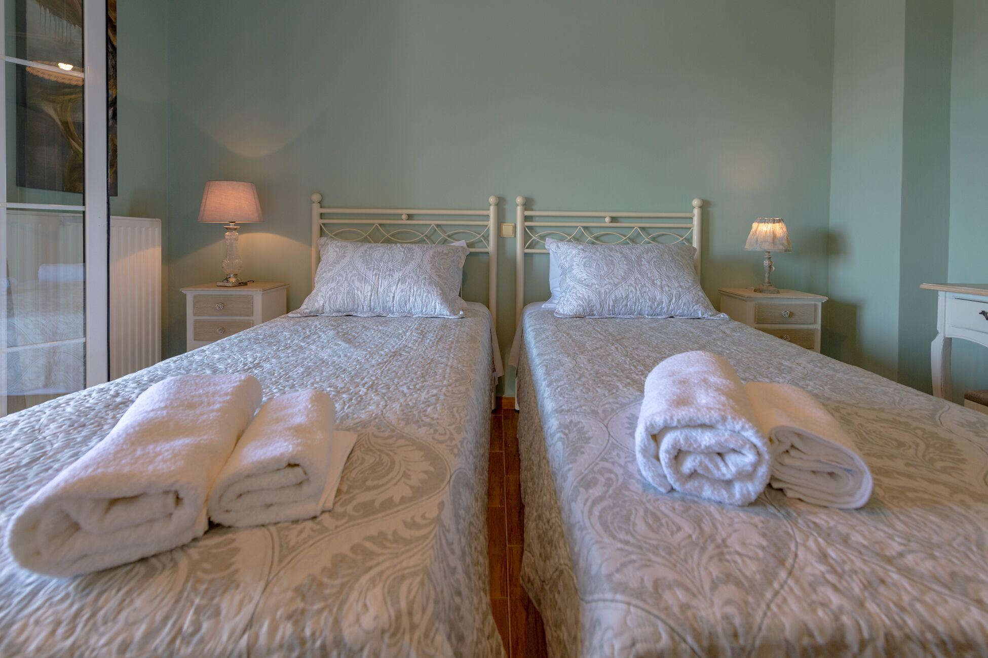 A hotel room with two beds, each with a white patterned bedspread and matching pillows. There are two bedside tables with lamps, and the room has a simple, clean design.