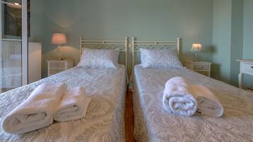 A hotel room with two beds, each with a white patterned bedspread and matching pillows. There are two bedside tables with lamps, and the room has a simple, clean design.