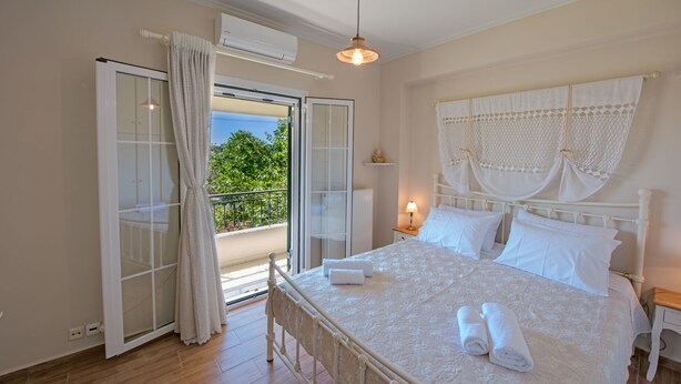 A bedroom with a bed, a nightstand, a lamp, a window with curtains, and an air conditioning unit. - It is our pleasure to welcome you to Kefalonia Island and invite you to enjoy yo (kefalonia)
