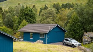 Une cabane bleue avec une voiture garée devant, entourée d’une pelouse et d’arbres.