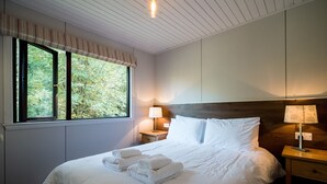 Un lit bien fait, avec du linge de lit et des serviettes blanches, une table de chevet en bois avec une lampe, et une fenêtre donnant sur des arbres.