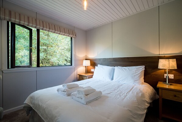Una cama bien hecha con sábanas y toallas blancas, una mesita de noche de madera con una lámpara, y una ventana con vistas a árboles.
