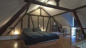 A bedroom with a sloped ceiling, exposed wooden beams, a bed with a dark cover, a small table with a chair, and a window allowing natural light. - Modern 5-star luxury in a historic setting (Boppard)