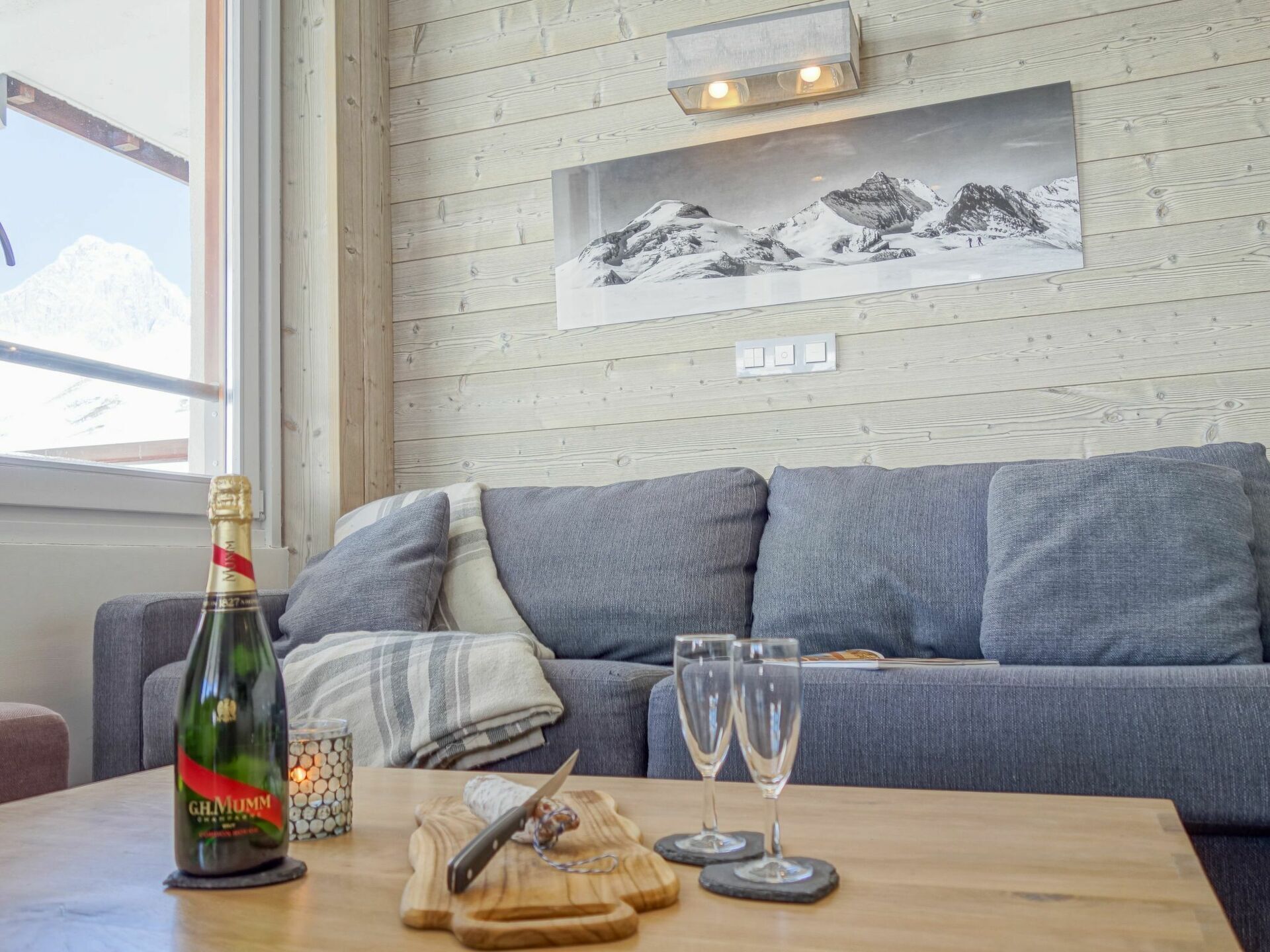 Ett mysigt vardagsrum med en grå soffa, ett träbord med en flaska champagne, två glas och en kniv på en skärbräda.