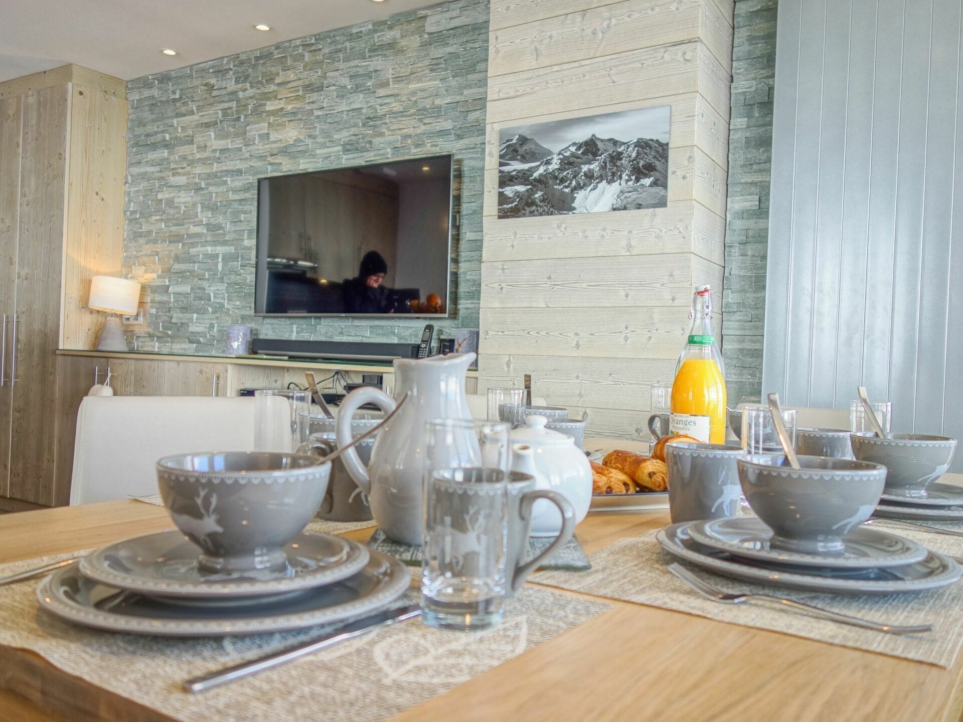 Un coin repas agrémenté d’un mur en pierre, d’un téléviseur à écran plat et d’un buffet pour le petit-déjeuner comprenant des croissants, du jus d’orange et des tasses à thé.