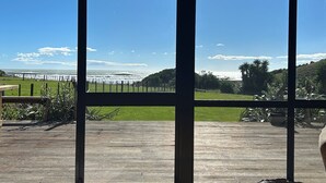 A view through large windows of a wooden deck overlooking a grassy area with a fence and a body of water. - 'Time Out'  Beach Front getaway with amazing sea views (Whangaehu Beach)