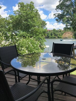 Ein runder Glastisch mit Korbstühlen auf einer Holzterrasse mit Blick auf einen See und Boote.