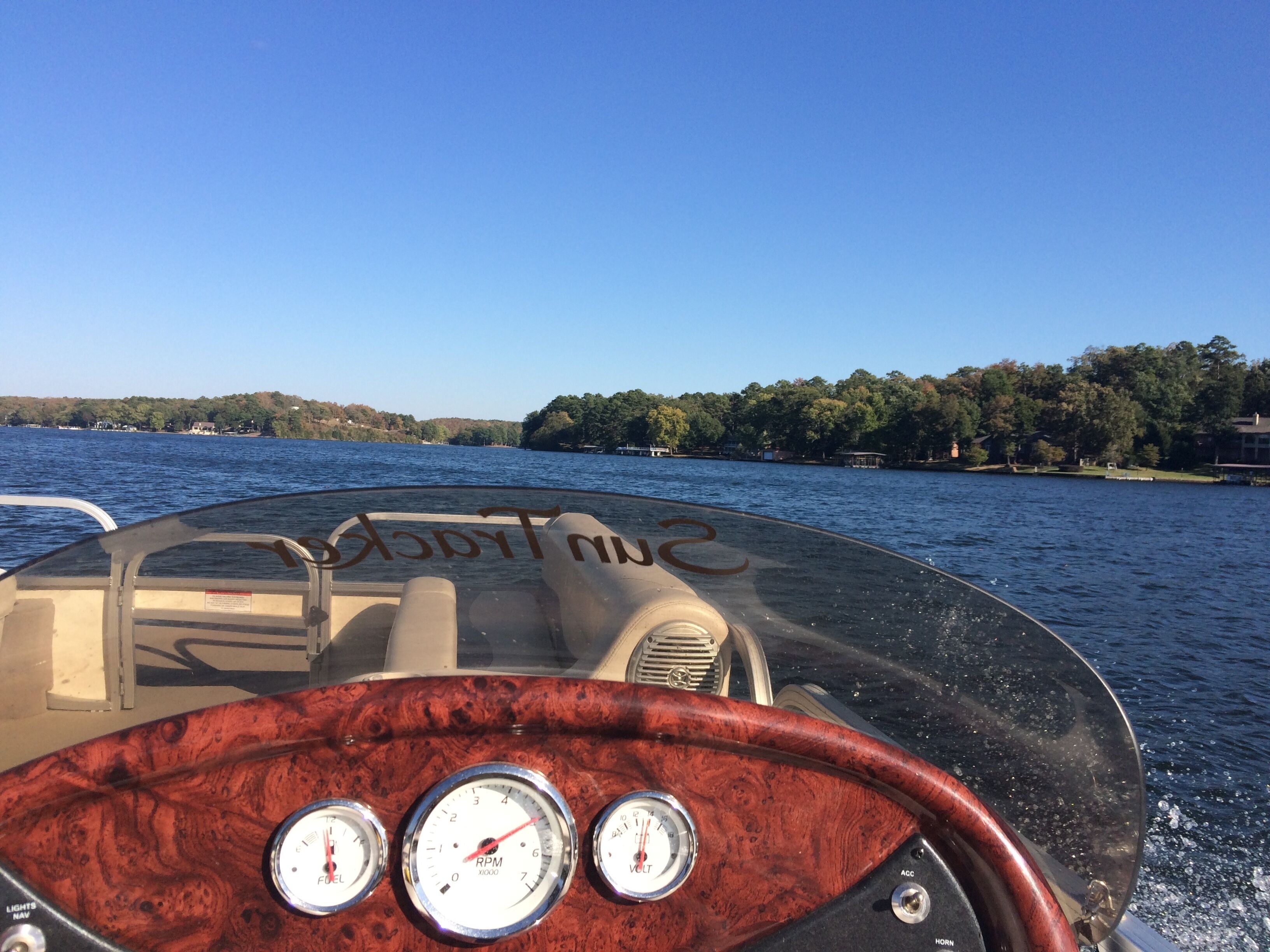 A boat's steering wheel with gauges and a clear view of the water and trees in the background.