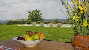 Une table avec un saladier de fruits et un vase de fleurs, une piscine et une zone herbeuse avec un arbre et des montagnes en arrière-plan.