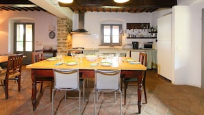 Un coin repas avec une grande table en bois, dressée pour six personnes, entourée de chaises, dans une cuisine rustique aux poutres apparentes et au mur en pierre.
