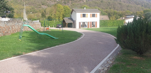 A paved driveway curving through a residential area with a house, a swing set, and a car parked nearby. - House of 100 m2 on a 2500 m2 fenced (Cierp-Gaud)