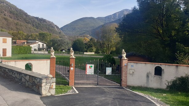 A gated entrance to a property with a stone wall, a paved driveway, and a view of a green landscape with trees and mountains in the background. - House of 100 m2 on a 2500 m2 fenced (Cierp-Gaud)