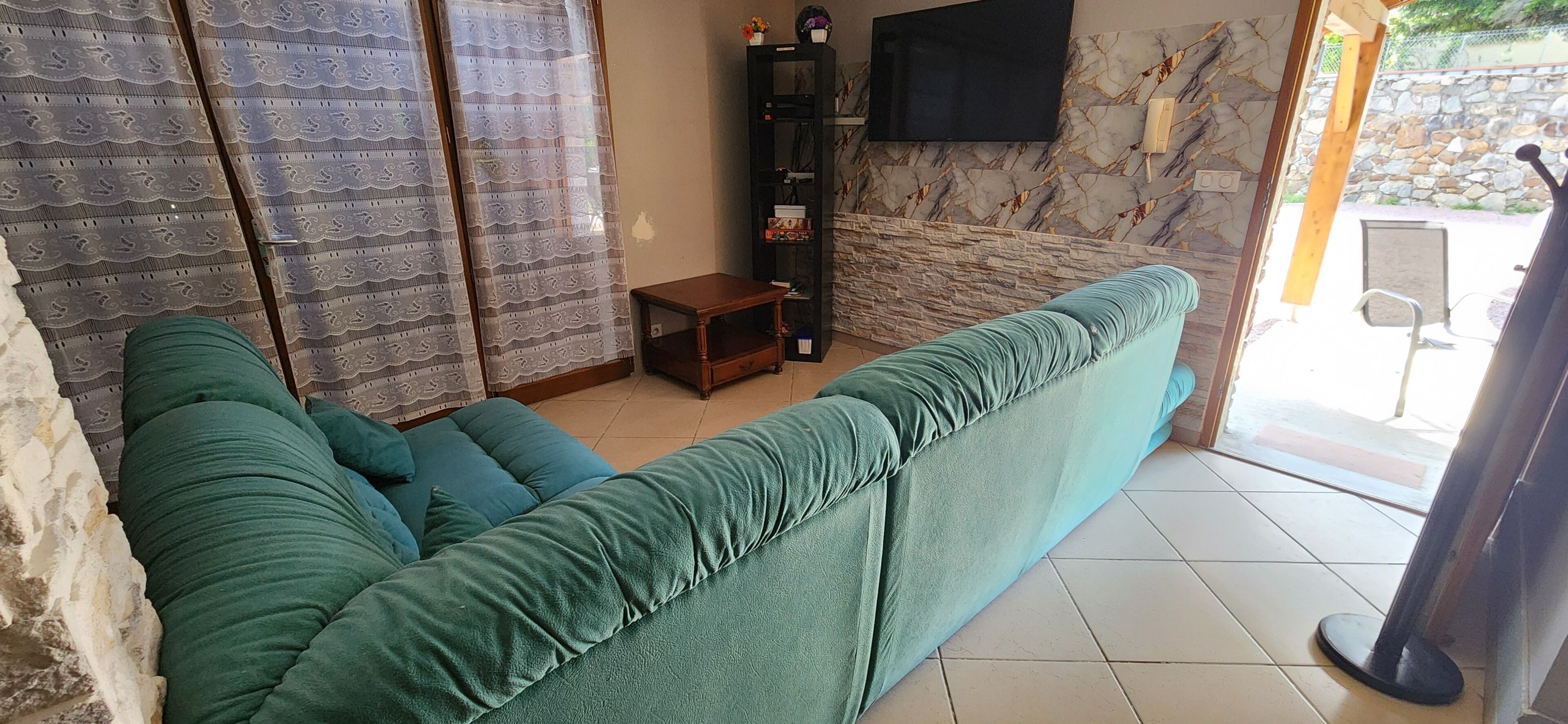 Un salon avec un canapé vert, une petite table d’appoint en bois, un téléviseur fixé sur un mur en pierre, et un mur en pierre à l’extérieur.