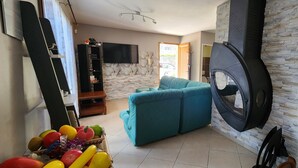 A living room with a sofa, a flat-screen TV, a stone wall, and a kitchen area. - House of 100 m2 on a 2500 m2 fenced (Cierp-Gaud)
