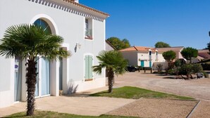 A white building with a tiled roof, a door with a green shutter, and a small balcony. There are palm trees in front of the building. - House for 4, close to the beach, shared pool (Saint-Hilaire-de-Riez)