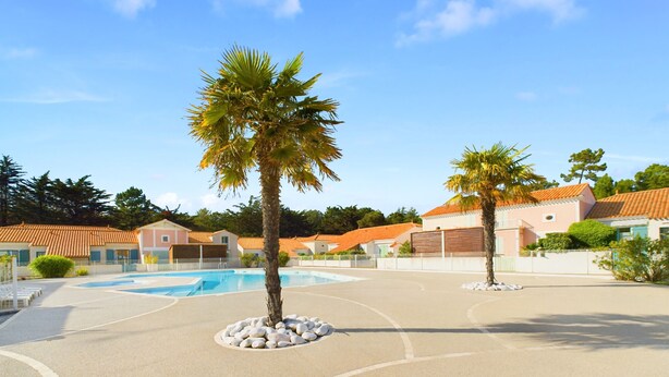 A pool area with palm trees, a clear sky, and buildings with orange roofs. - House for 4, close to the beach, shared pool (Saint-Hilaire-de-Riez)