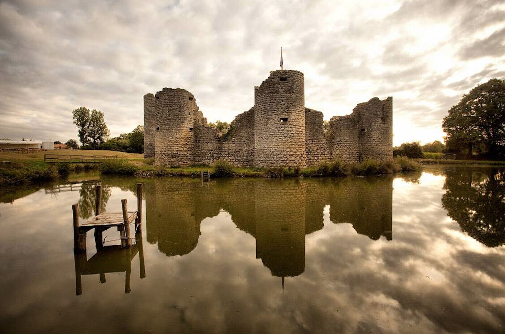 A historic stone castle with a moat, a wooden dock, and a clear reflection in the water.