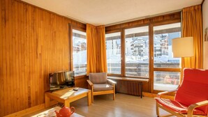 Ein gemütliches Zimmer mit Holzwänden, einem Fernseher, einem roten Sessel, einem grauen Sessel, einem Holztisch mit Teekanne und Tassen sowie großen Fenstern mit Vorhängen.