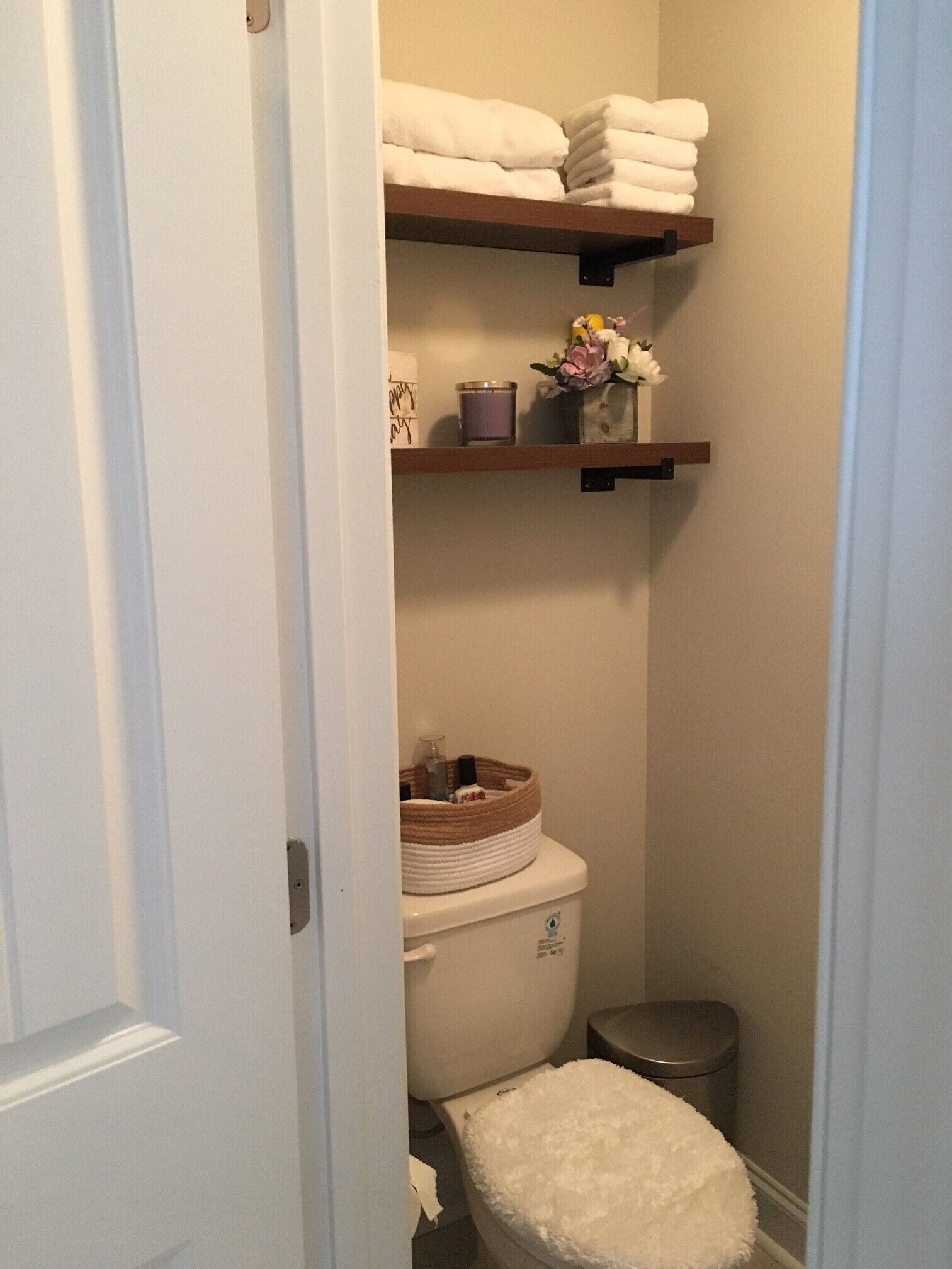 A compact bathroom with a toilet, a small trash bin, and shelves holding towels and toiletries.