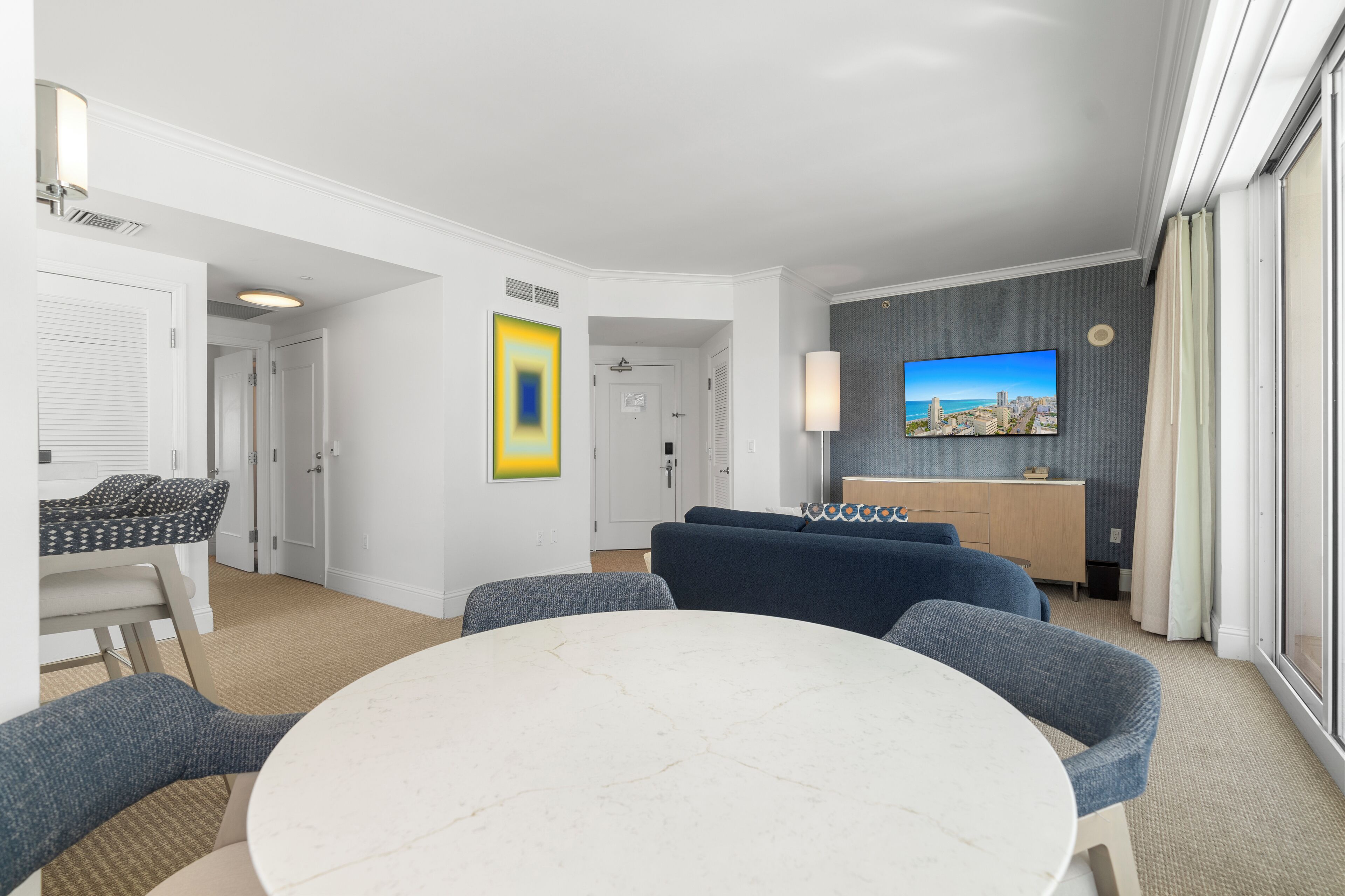 A modern hotel room with a round dining table, blue upholstered seating, a kitchenette, and a large window with curtains.