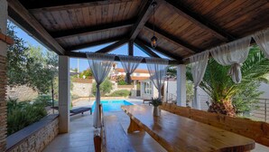 A covered outdoor seating area with wooden benches and a pool in the background. - Villa Parlera (Ližnjan)