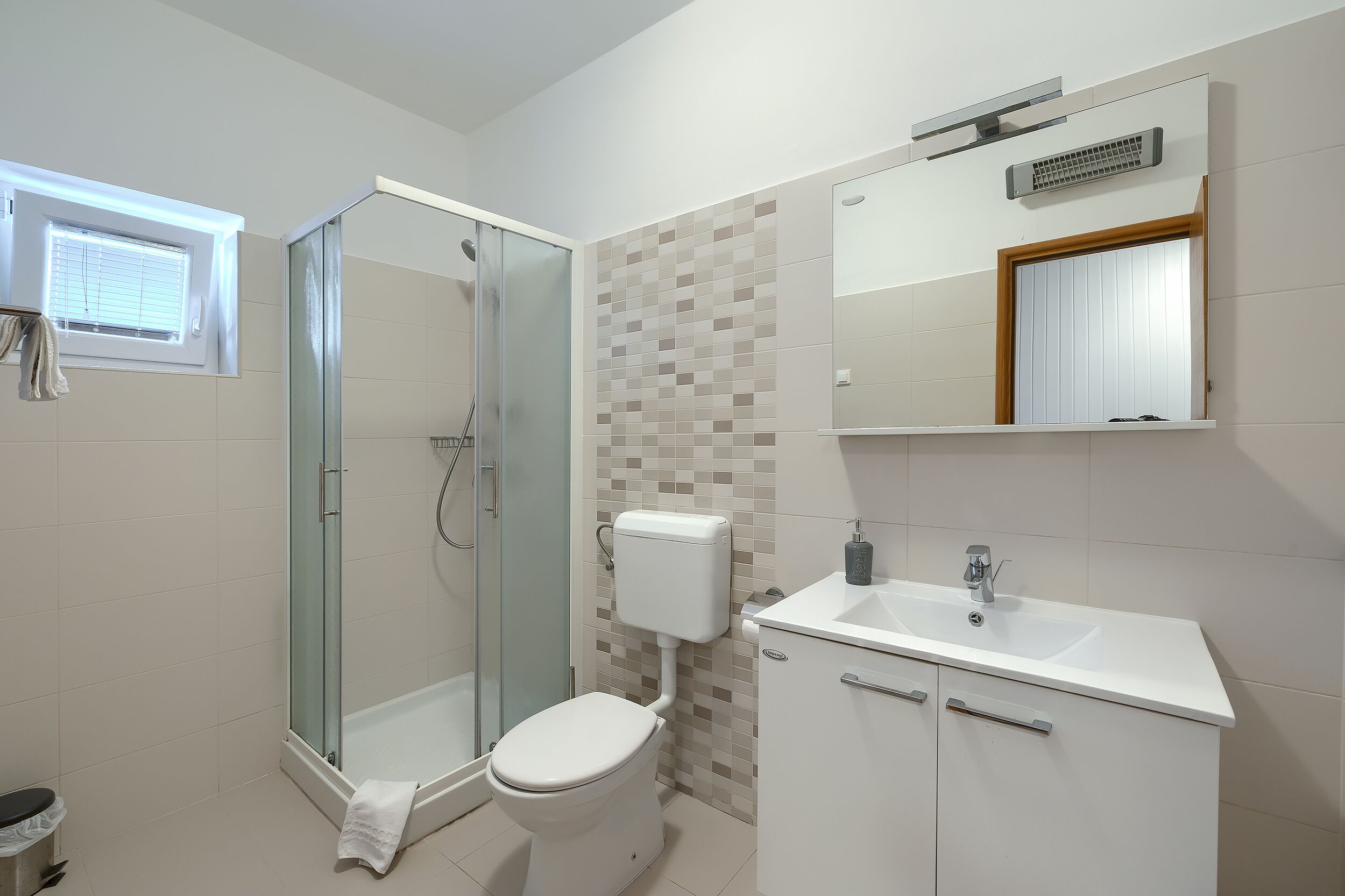 Une salle de bain moderne avec une douche en verre, des toilettes, un lavabo avec un miroir et une fenêtre avec des stores.
