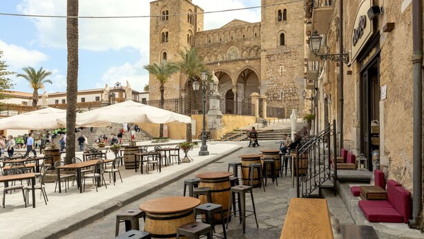 Un área de café al aire libre con mesas y sillas, una palmera y un edificio histórico con una torre al fondo.
