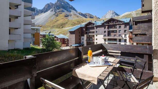 Un espace extérieur aménagé en bois, avec une table et des bancs, offrant une vue sur une chaîne de montagnes et des bâtiments aux façades en bois.