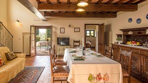 Un coin repas avec une longue table dressée pour six personnes, des chaises en bois, un téléviseur et une vue sur l’extérieur.