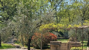 Un sendero en un jardín con pavimento de piedra, rodeado de árboles y arbustos diversos, que conduce a una estructura de pérgola.