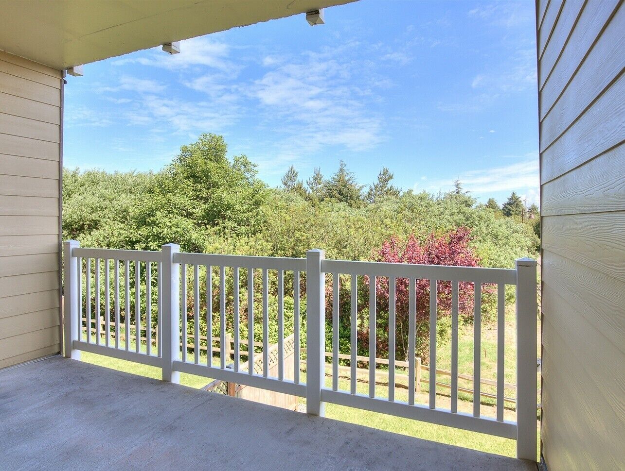 A covered porch with a white fence, a concrete floor, and a view of trees and shrubs.